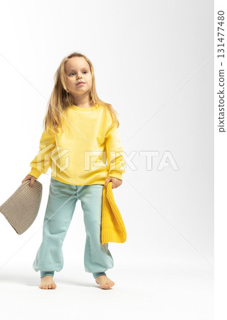 Portrait of beautiful cute little toddler girl. Child with funny face in yellow sweatshirt plays with hats. Pretty smile kid in studio on white background. High quality photo Portrait of beautiful cute little toddler girl. Child with funny face in yellow sweatshirt plays with hats. Pretty smile kid in studio on white background. High quality photo 131477480