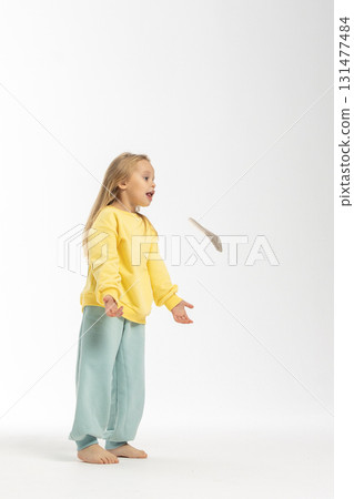 Portrait of beautiful cute little toddler girl. Child with funny face in yellow sweatshirt plays with hats. Pretty smile kid in studio on white background. High quality photo 131477484