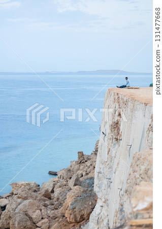 Man greets sunrise of sun on high mountain above sea. Aerial view to calm blue sea with rocky coastline. Panoramic view of dawn above horizont in summer morning, Sharm El Sheikh, Egypt.  131477668