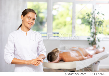 Smiling young female masseur in white coat standing in massage room Smiling young female masseur in white coat standing in massage room 131477669
