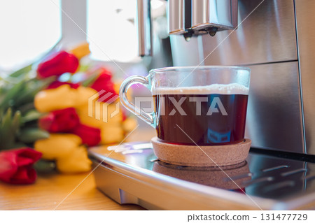 Cup of coffee with milk and tulips flowers on wooden kitchen table. Freshly brewed cappuccino espresso in coffee machine for breakfast. Stylish home interior decor. High quality photo 131477729