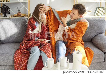 Two sick young adults sitting on sofa under blankets, supporting each other during illness at home. Two sick young adults sitting on sofa under blankets, supporting each other during illness at home. 131478102