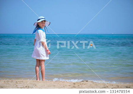 Rear view of child girl in white summer dress and sunshade hat with blue ribbons in long braids standing barefoot on sandy beach enjoying sea view 131478554