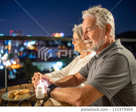 AI generated image of an elderly couple sitting on a balcony at night. The man smartwatch alerts him to take his medication. The peaceful city lights and modern setting create a calm atmosphere. 131479968