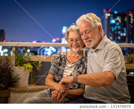 AI generated image of an elderly couple sitting on a balcony at night. The man smartwatch alerts him to take his medication. The peaceful city lights and modern setting create a calm atmosphere. 131479970
