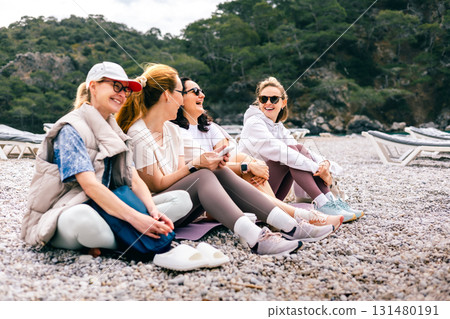 Group of smiling women relaxing on a pebbly beach in sportswear, enjoying outdoor conversation 131480191