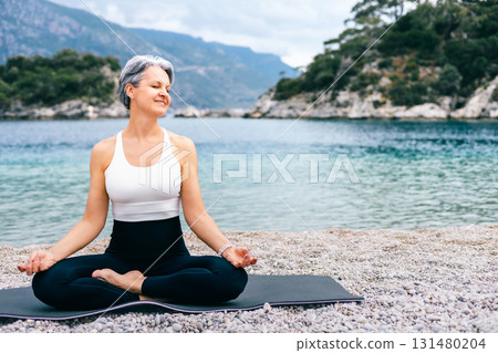 Smiling senior woman practicing yoga meditation at the seaside with scenic mountain view Smiling senior woman practicing yoga meditation at the seaside with scenic mountain view 131480204