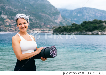 Smiling senior woman holding yoga mat on beach, embracing active and healthy aging outdoors 131480319