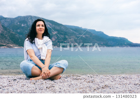Relaxed Woman Sitting Cross-Legged on Pebble Beach by the Sea with Mountain View Behind 131480323