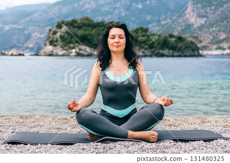 Young woman meditating in lotus pose on rocky beach with peaceful sea and mountain backdrop 131480325