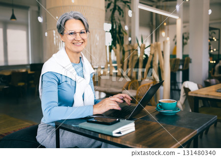 Smiling senior freelance woman with gray hair working on laptop in cafe, confident and independent 131480340