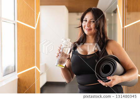 Smiling woman with yoga mat and water bottle standing in bright modern fitness center hallway Smiling woman with yoga mat and water bottle standing in bright modern fitness center hallway 131480345