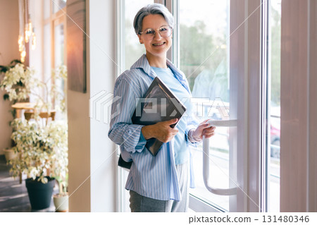 Mature woman holding laptop and smiling while opening modern glass door in cozy indoor setting Mature woman holding laptop and smiling while opening modern glass door in cozy indoor setting 131480346