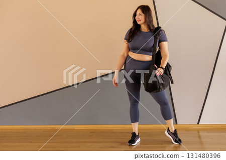 Full figured woman in activewear carrying gym bag in bright studio after yoga workout Full figured woman in activewear carrying gym bag in bright studio after yoga workout 131480396