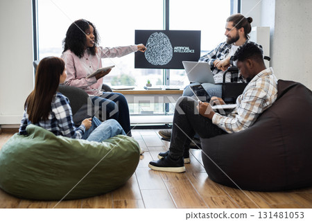Group of men and women collaborating on artificial intelligence presentation in relaxed office. Includes professionals with diverse backgrounds, surrounded by beanbags, using digital technology 131481053