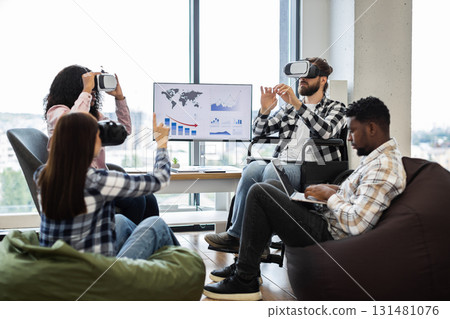 Diverse group using VR glasses and laptops in teamwork collaboration in office environment. Interacting with technology while seated on bean bags and chairs, showcasing inclusivity 131481076