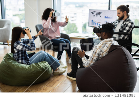 Group of diverse adults using virtual reality headsets and laptops in modern office setting. Individuals of various backgrounds collaborating in technology-driven workspace fostering creativity 131481095