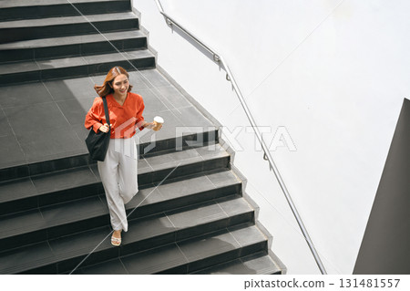 Confident young woman walking down stairs, holding coffee cup and bag, concept of modern lifestyle Confident young woman walking down stairs, holding coffee cup and bag, concept of modern lifestyle 131481557