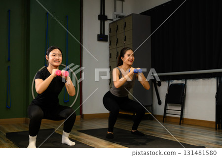 Female trainer and student working out together, holding weights and doing squats for muscle building and fitness Female trainer and student working out together, holding weights and doing squats for muscle building and fitness 131481580