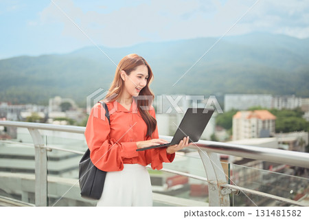 Happy businesswoman standing on balcony working on laptop, concept of digital nomad lifestyle 131481582