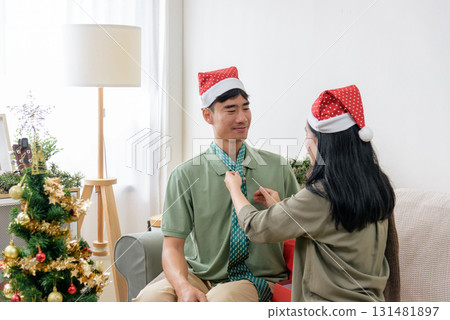 Christmas loving couple wearing festive santa hats celebrate holiday at home, winter joy fills festive living room, woman adjusts man tie near sparkling decorated christmas tree. 131481897