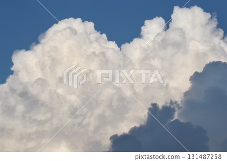 A majestic formation of towering, sunlit white cumulonimbus clouds against a deep blue sky, with dark grey shadows at the base, creating dramatic contrast and depth. A majestic formation of towering, sunlit white cumulonimbus clouds against a deep blue sky, with dark grey shadows at the base, creating dramatic contrast and depth. 131487258