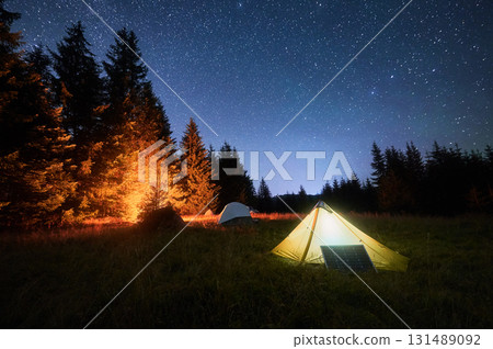 Yellow tent beside solar panel under stunning, star-filled night sky. Nearby, other tents and trees are lit by warm light from campfire, highlighting eco-friendly and serene camping experience. Yellow tent beside solar panel under stunning, star-filled night sky. Nearby, other tents and trees are lit by warm light from campfire, highlighting eco-friendly and serene camping experience. 131489092