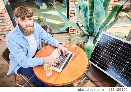 Man works on laptop at outdoor round table, with glass of iced coffee. Solar panel nearby, emphasizing sustainable, modern eco-friendly workspace that combines technology and sustainability. 131489101