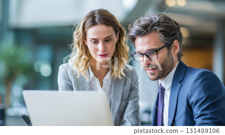 Two professional business people engrossed in a laptop screen within a modern office. 131489106