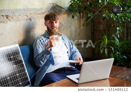 Bearded man works on laptop at wooden table, with cup of coffee. Solar panel nearby, emphasizing sustainable eco-friendly workspace that combines technology and sustainability in cozy environment. 131489112