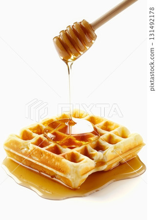 Golden honey pours down from a wooden dipper, covering a warm waffle surrounded by a glossy pool of syrup on a simple backdrop 131492178