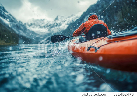 A person in a bright orange jacket paddles a kayak on a tranquil lake, with mountains looming in the background and rain splashing on the surface A person in a bright orange jacket paddles a kayak on a tranquil lake, with mountains looming in the background and rain splashing on the surface 131492476