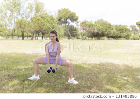 Woman exercising sumo squatting with dumbbells outdoors 131493886