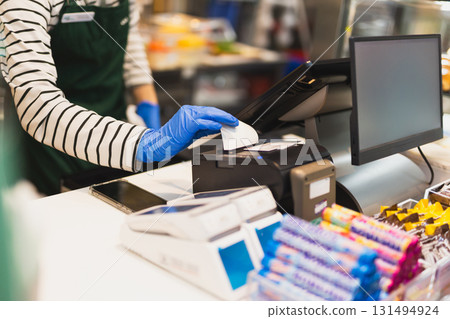 Woman cashier hand with blue grove taking receipt from cash register in a shop. Woman cashier hand with blue grove taking receipt from cash register in a shop. 131494924