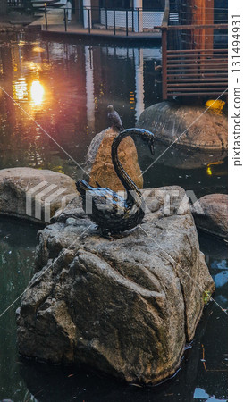 A serene Japanese garden featuring a tranquil lake surrounded by lush greenery and colorful autumn leaves. Sculptures are placed throughout the landscape. 131494931