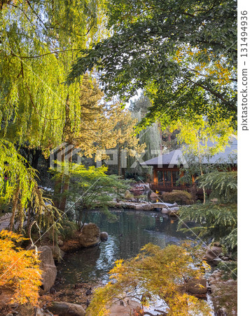 A serene Japanese garden featuring a tranquil lake surrounded by lush greenery and colorful autumn leaves. Sculptures are placed throughout the landscape. 131494936