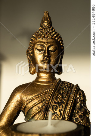 Golden Buddha statue holding a candle holder with a white candle, seated in meditation pose with shadow on beige background. Golden Buddha statue holding a candle holder with a white candle, seated in meditation pose with shadow on beige background. 131494996
