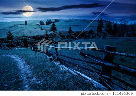 beautiful rural landscape in mountains at night in spring. road near the wooden fence through the grassy pasture with haystack and coniferous trees on hillside in full moon light 131495369