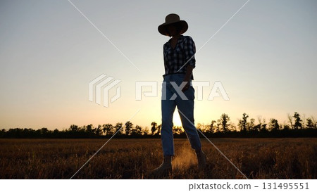 Adult agronomist in straw hat walking among barley plantation at dusk. Female farmer going through wheat meadow at sunset. Beautiful scenic landscape. Concept of agricultural business. Slow motion 131495551