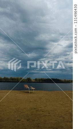 A sun lounger stands on a sandy beach by the river. The beach landscape features a beautiful sky. Dark clouds are in the sky. 131496430