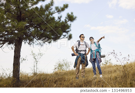 Hiking couple with backpacks exploring hillside meadow, pointing at forest view 131496953