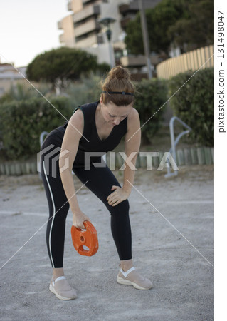 Woman exercising outdoors, holding weight disc, feeling tired Woman exercising outdoors, holding weight disc, feeling tired 131498047
