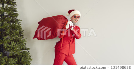Stylish middle-aged Santa Claus carrying heavy red sack full of Christmas presents on his shoulders Stylish middle-aged Santa Claus carrying heavy red sack full of Christmas presents on his shoulders 131498658