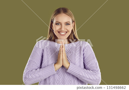 Grateful smiling young blondy woman in purple sweater folded hands in prayer on khaki background. 131498662