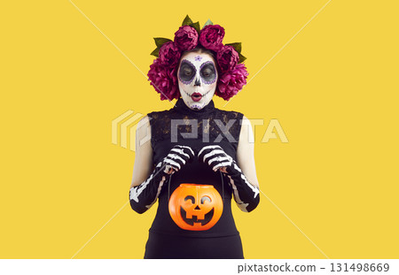 Woman in Halloween costume with wreath of pink flowers and scary make-up holding a pumpkin lantern. Woman in Halloween costume with wreath of pink flowers and scary make-up holding a pumpkin lantern. 131498669