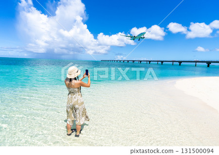 一位身穿洋裝的年輕女子用智慧型手機拍攝一架飛機降落在翡翠海灘上的照片 131500094