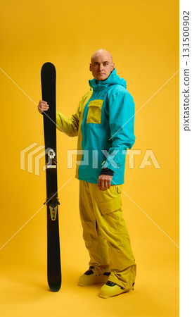 Skier standing with ski held upright on bright yellow studio background 131500902