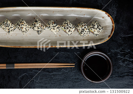 A row of sushi rolls, coated in a mix of black and white sesame seeds, is elegantly arranged on a long ceramic plate. A row of sushi rolls, coated in a mix of black and white sesame seeds, is elegantly arranged on a long ceramic plate. 131500922