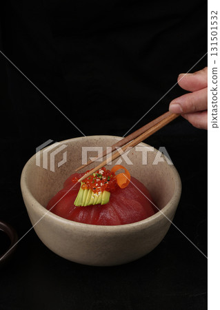 A delicate tuna dome topped with avocado slices, salmon roe, and carrot ribbons, served in a ceramic bowl, captured with a hand holding chopsticks for an elegant touch. 131501532
