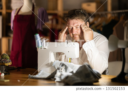 Overworked middle-aged male tailor sitting on table next to sewing machine 131502153